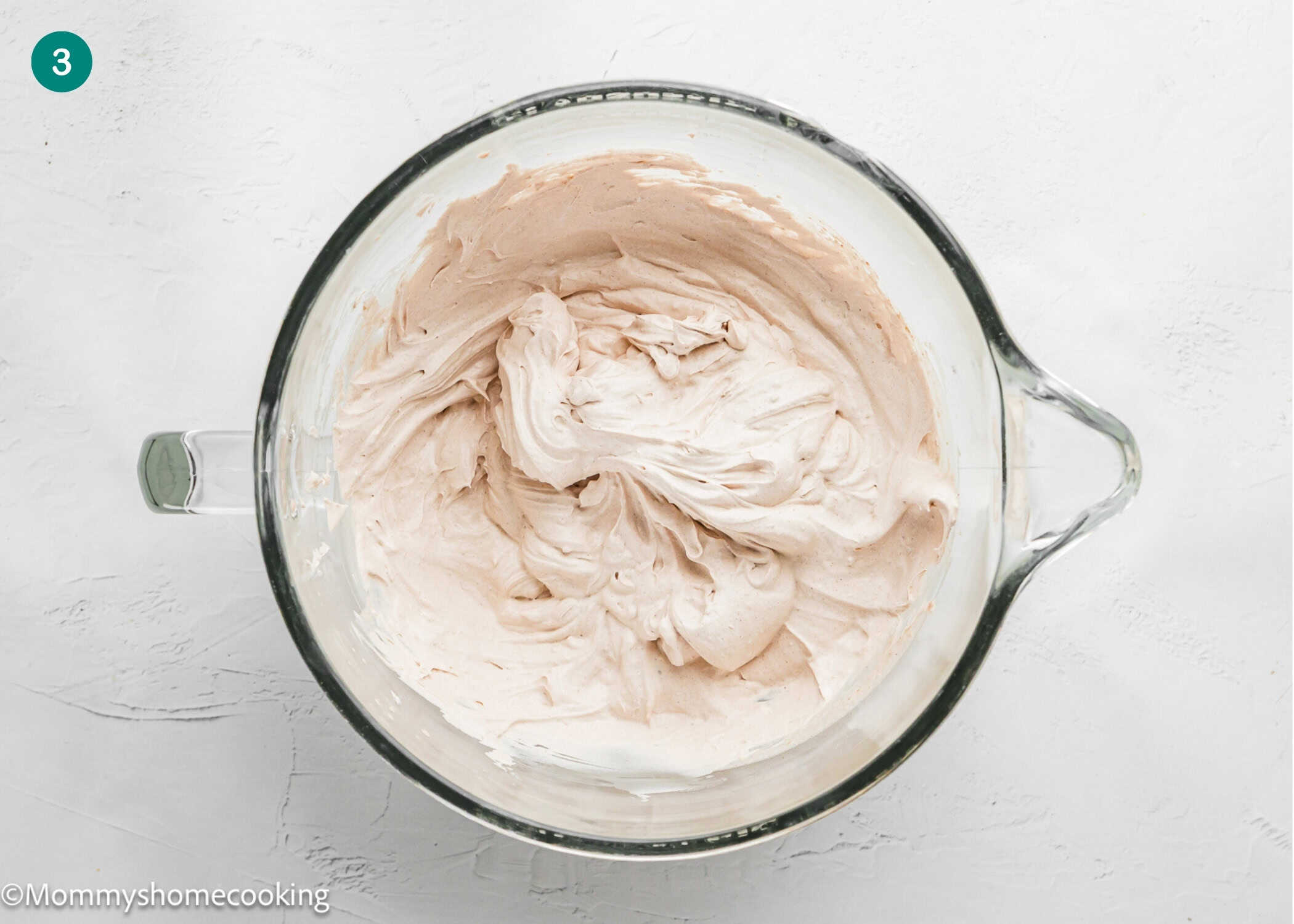 A glass mixing bowl filled with light brown whipped frosting for an Eggless Yule Log (Bûche De Noël) sits on a white surface.