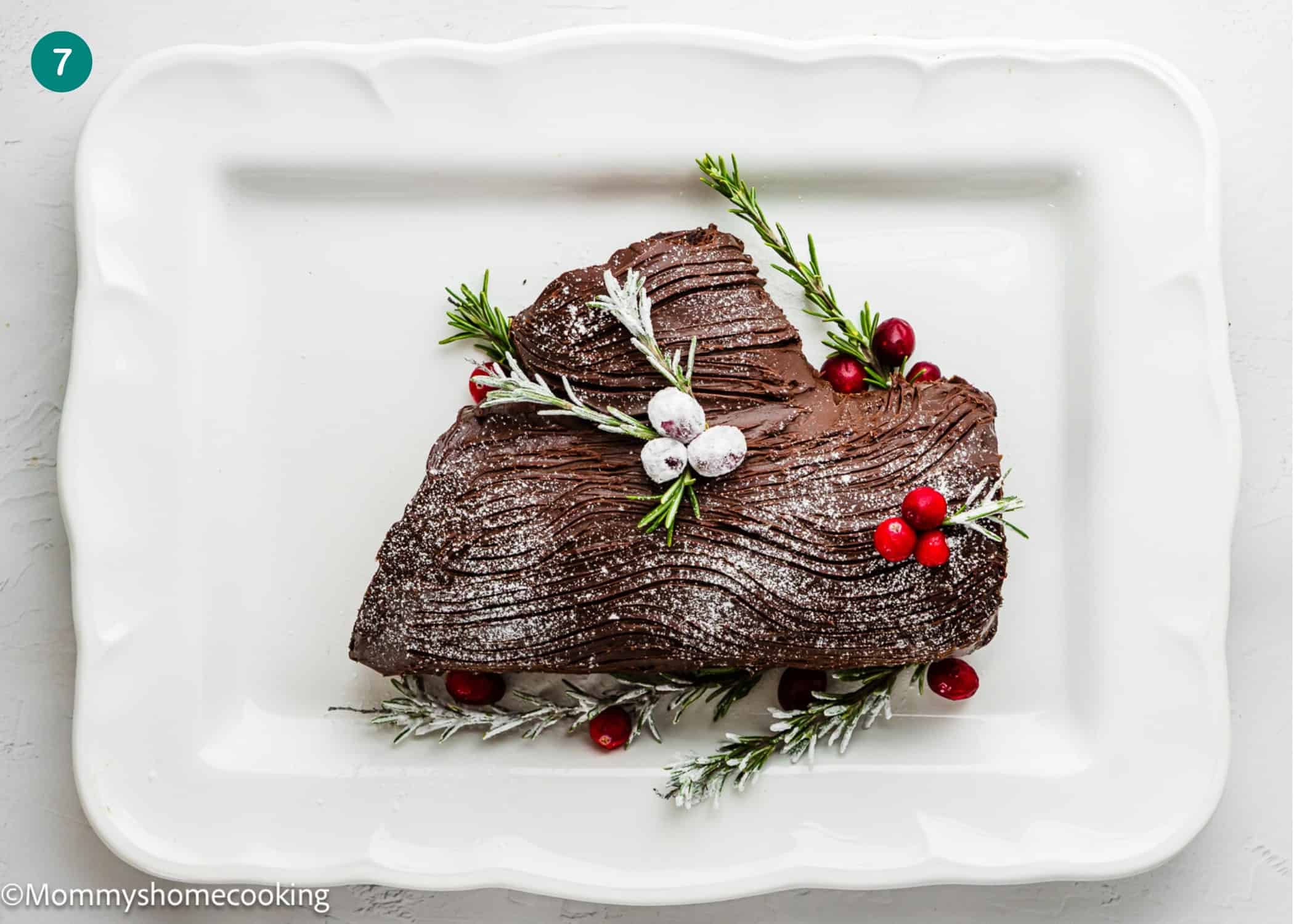 An Eggless Yule Log (Bûche De Noël) cake decorated with frosted cranberries, rosemary, and powdered sugar, served on a white rectangular plate.