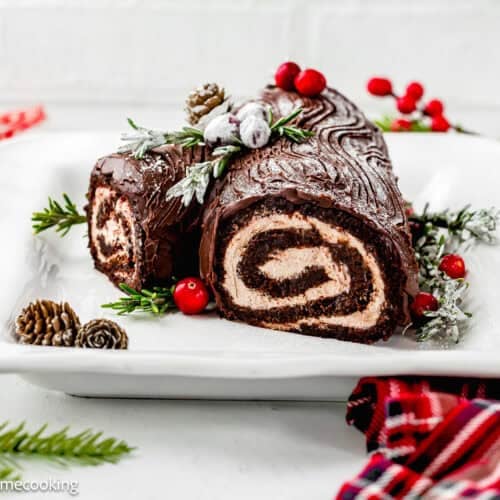 An Egg-Free Yule Log (Bûche De Noël) with cream filling is decorated with sugared cranberries, pine cones, and greenery on a white plate.