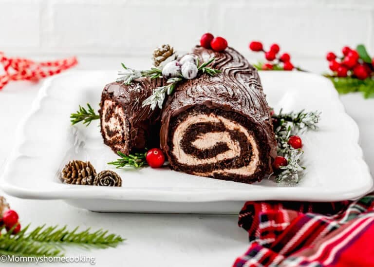 An Egg-Free Yule Log (Bûche De Noël) with cream filling is decorated with sugared cranberries, pine cones, and greenery on a white plate.