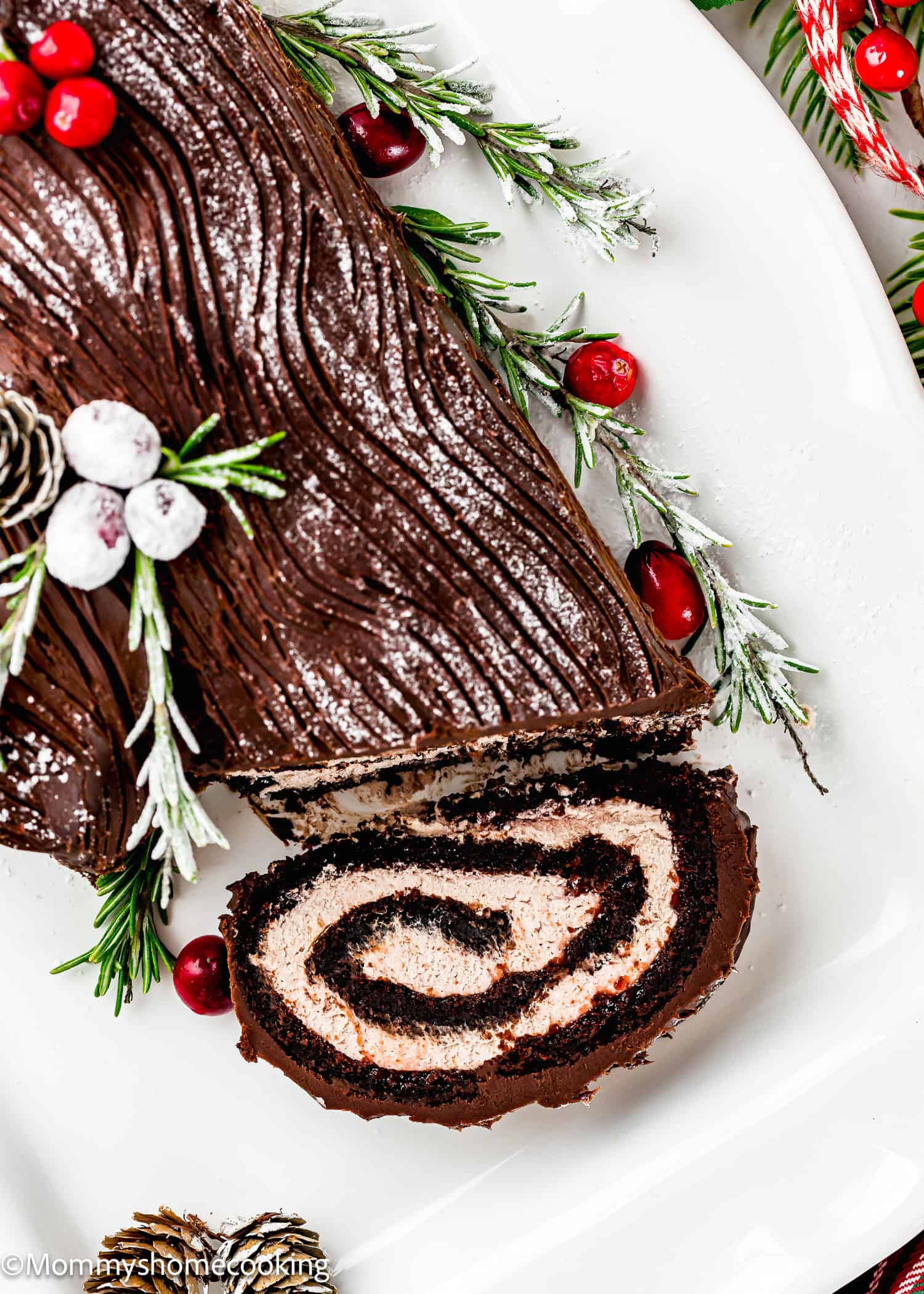 An Egg-Free Yule Log (Bûche De Noël) chocolate cake with a spiral filling is decorated with sugared cranberries, rosemary sprigs, and pinecones on a white plate.