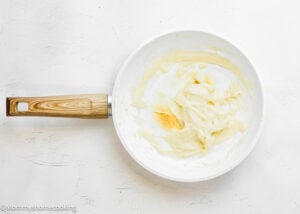 White nonstick pan with a wooden handle containing a smooth Tangzhong for Overnight Cinnamon Rolls partially spread in the center, resting on a white textured surface.