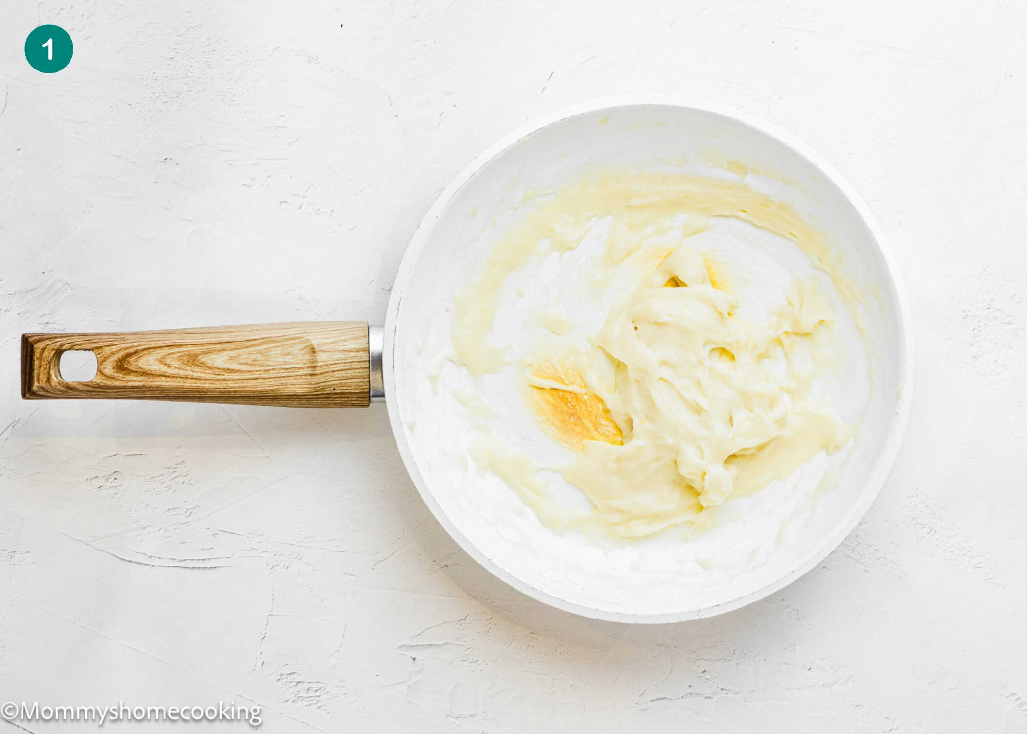 White nonstick pan with a wooden handle containing a smooth, pale batter for Overnight Cinnamon Rolls (Small Batch), partially spread in the center, resting on a white textured surface.