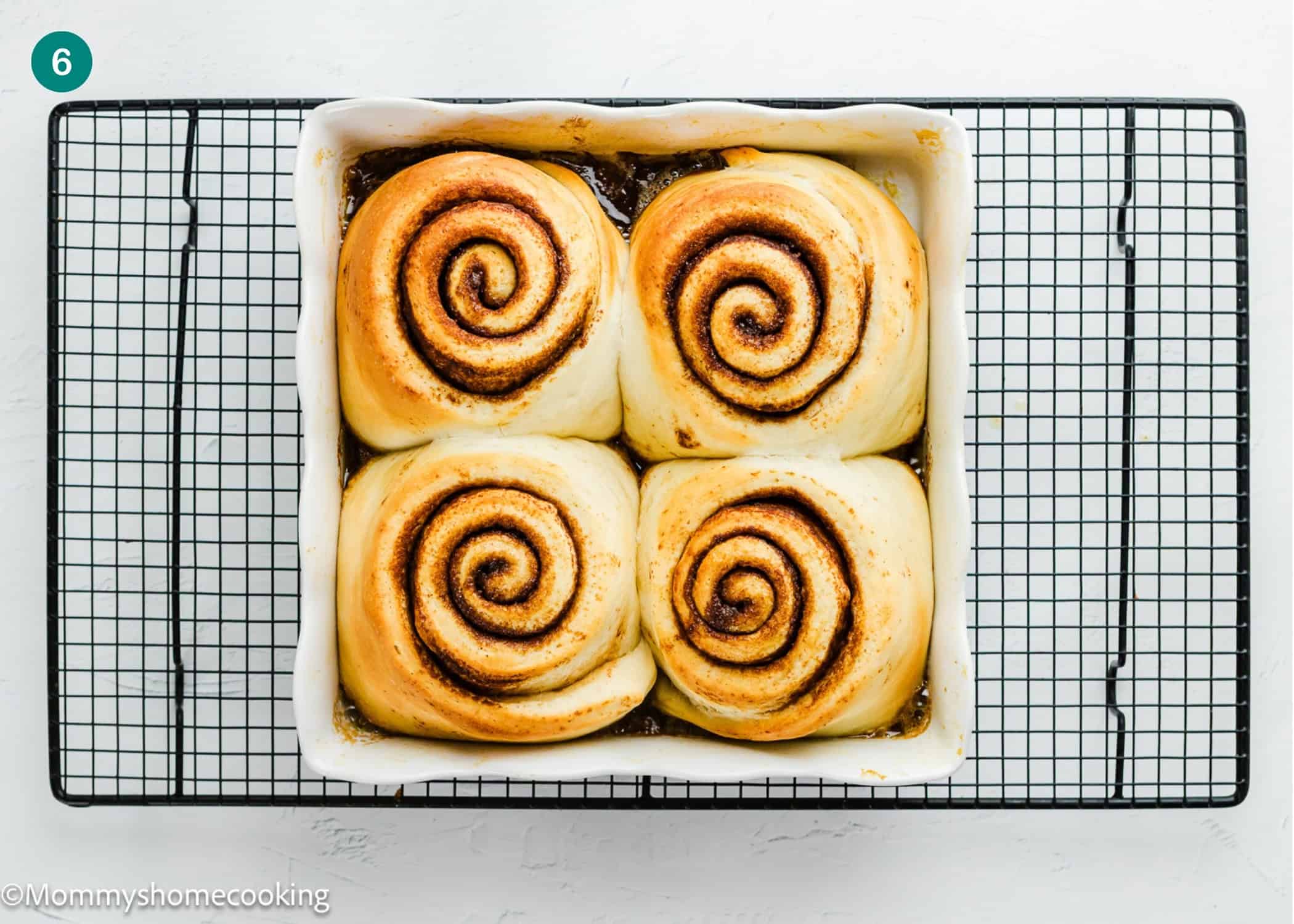 Four large Overnight Cinnamon Rolls (Small Batch) in a square white baking dish, resting on a black cooling rack over a white surface.