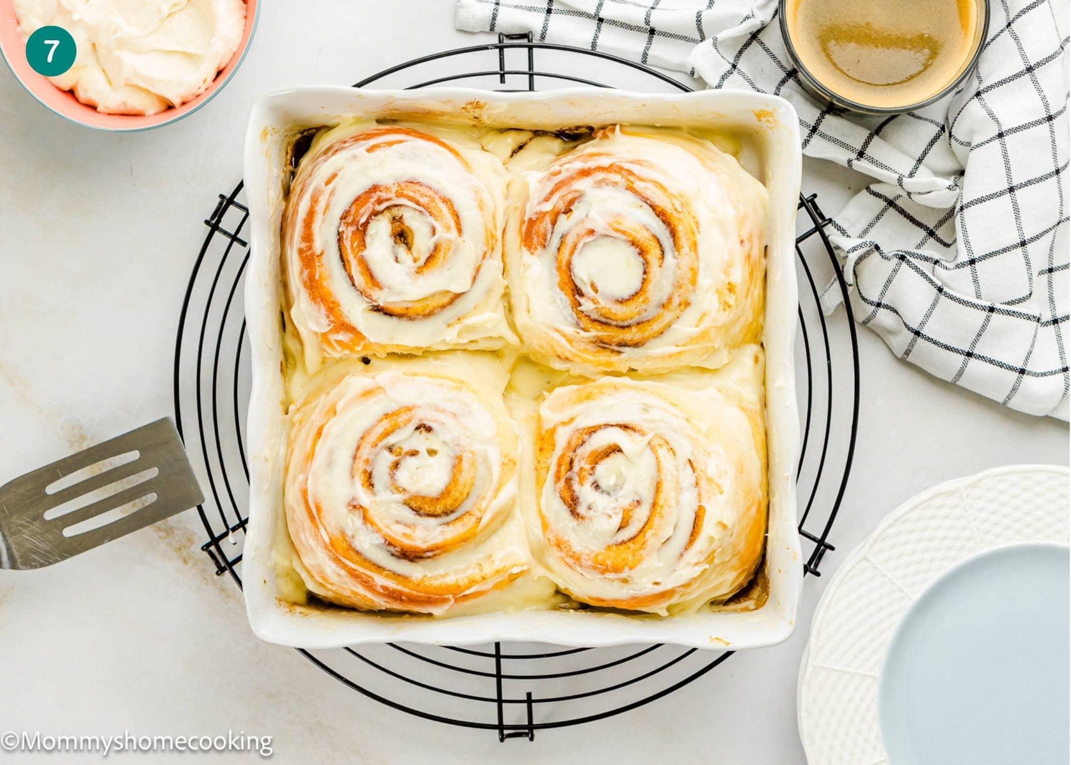 A baking dish with four Overnight Cinnamon Rolls (Small Batch) sits on a round cooling rack, surrounded by a spatula, a cup, a napkin, and a plate.