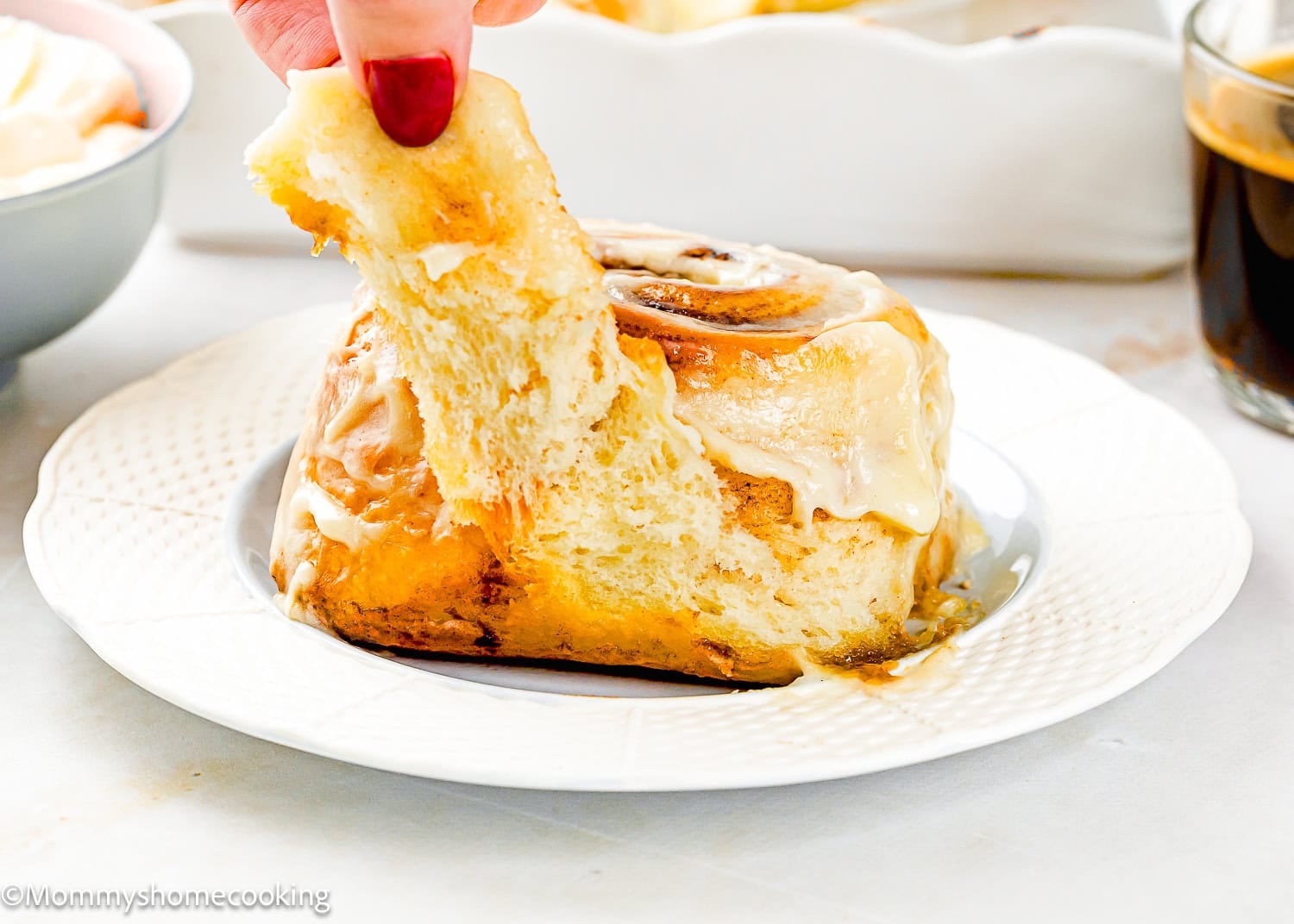 A hand pulls apart an Overnight Cinnamon Roll (Small Batch) topped with icing on a white plate, with a cup of coffee and a bowl in the background.