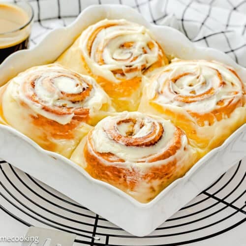Four large Overnight Cinnamon Rolls (Small Batch) in a white baking dish on a cooling rack, with a cup of coffee and a checked towel in the background.
