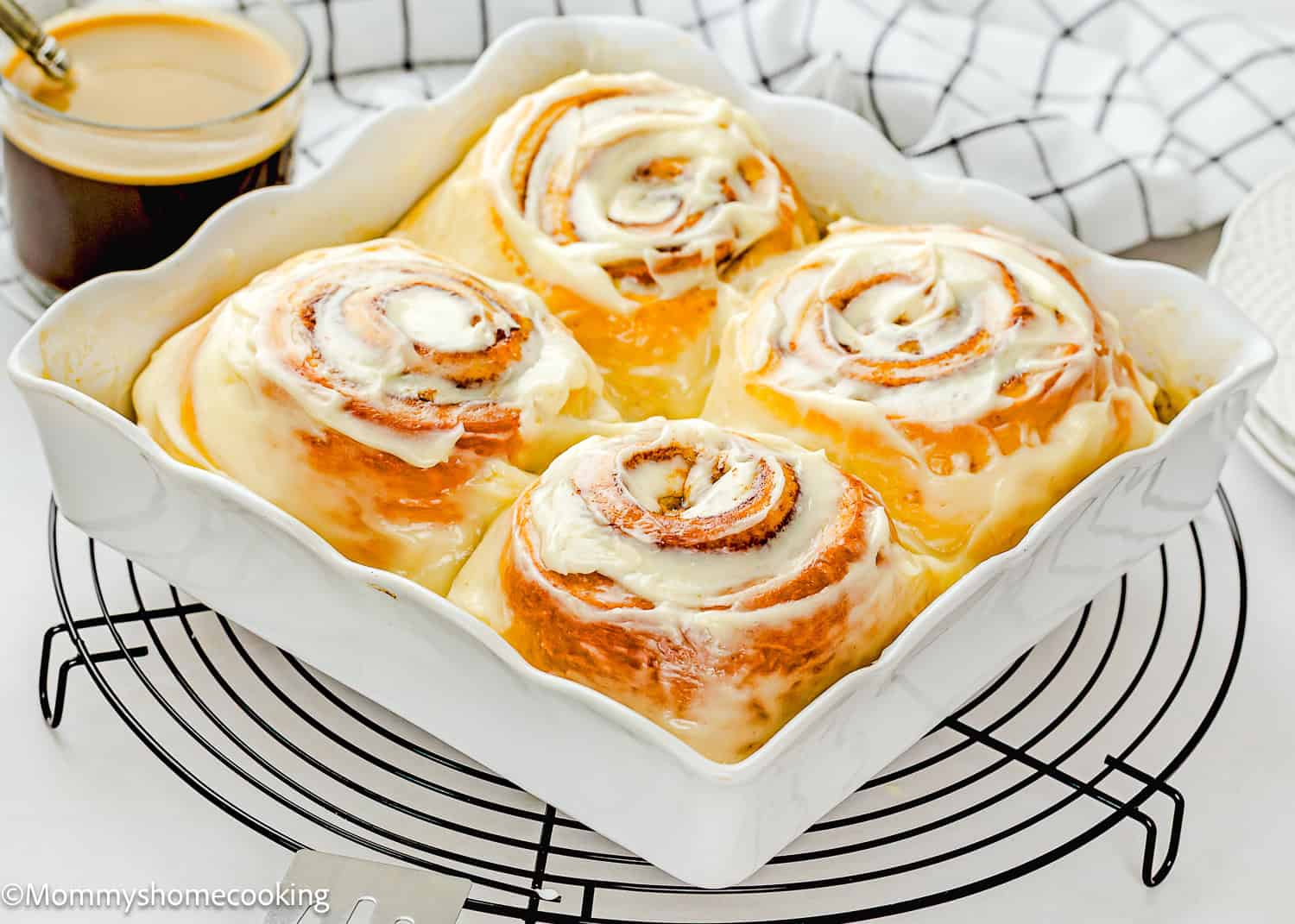 Four large Overnight Cinnamon Rolls (Small Batch) in a white baking dish on a cooling rack, with a cup of coffee and a checked towel in the background.