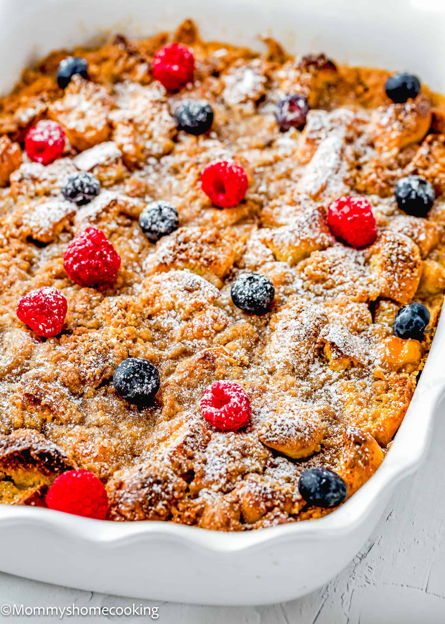 An Overnight French Toast Casserole Without Eggs, topped with powdered sugar, raspberries, and blueberries in a white ceramic pan.