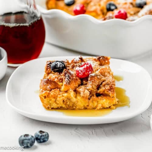 A slice of Overnight French Toast Casserole Without Eggs topped with berries and syrup sits on a white plate, surrounded by bowls of blueberries, raspberries, and a jar of syrup.
