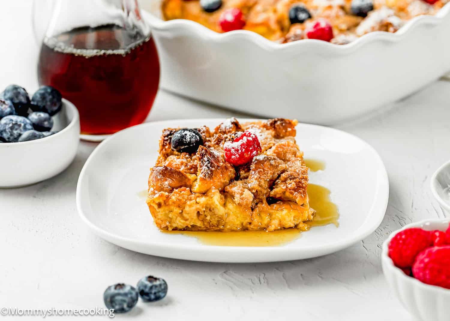 A slice of Overnight French Toast Casserole Without Eggs topped with berries and syrup sits on a white plate, surrounded by bowls of blueberries, raspberries, and a jar of syrup.