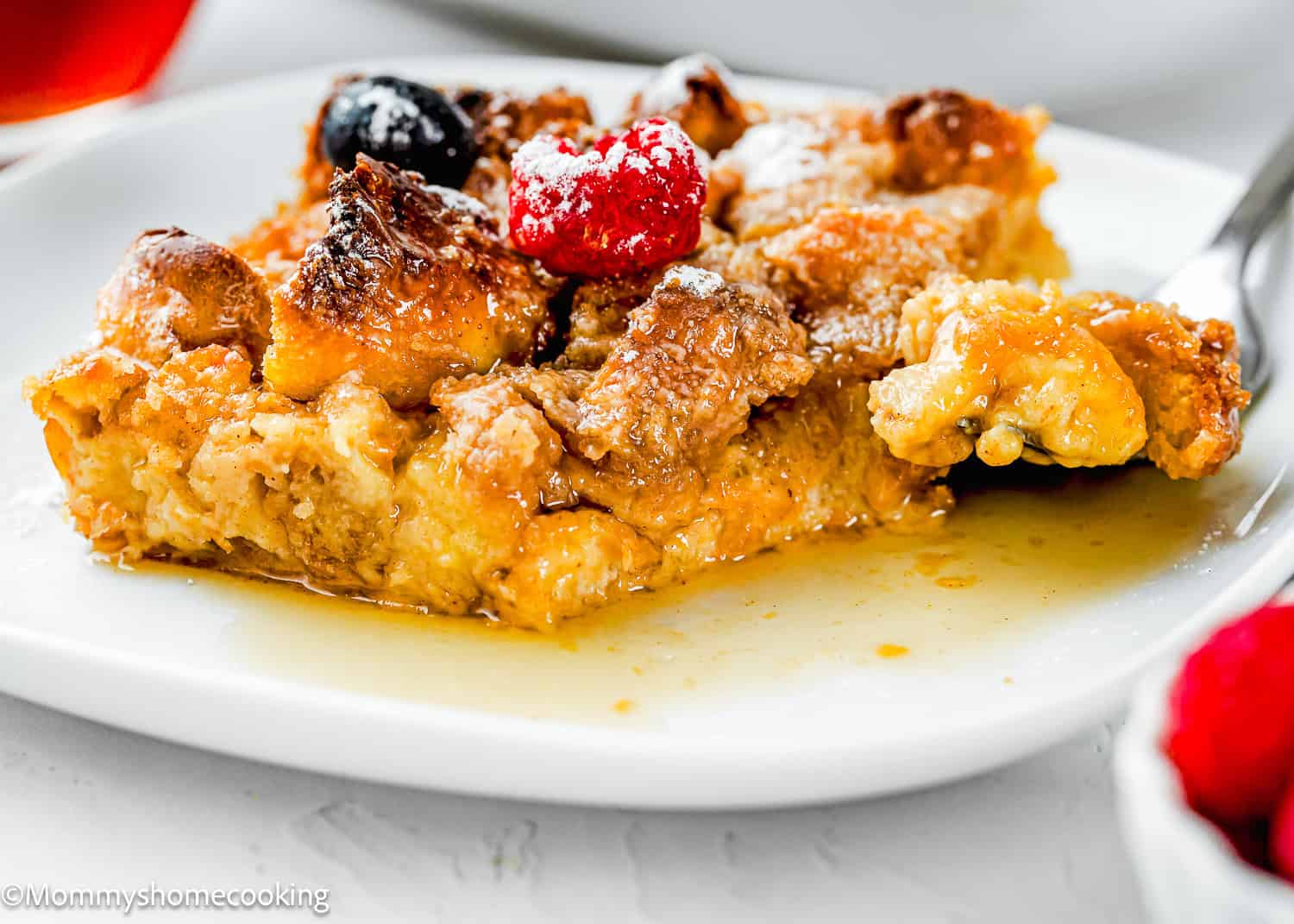 A slice of Overnight French Toast Casserole Without Eggs topped with berries sits on a white plate, with a fork taking a bite and syrup pooling underneath.