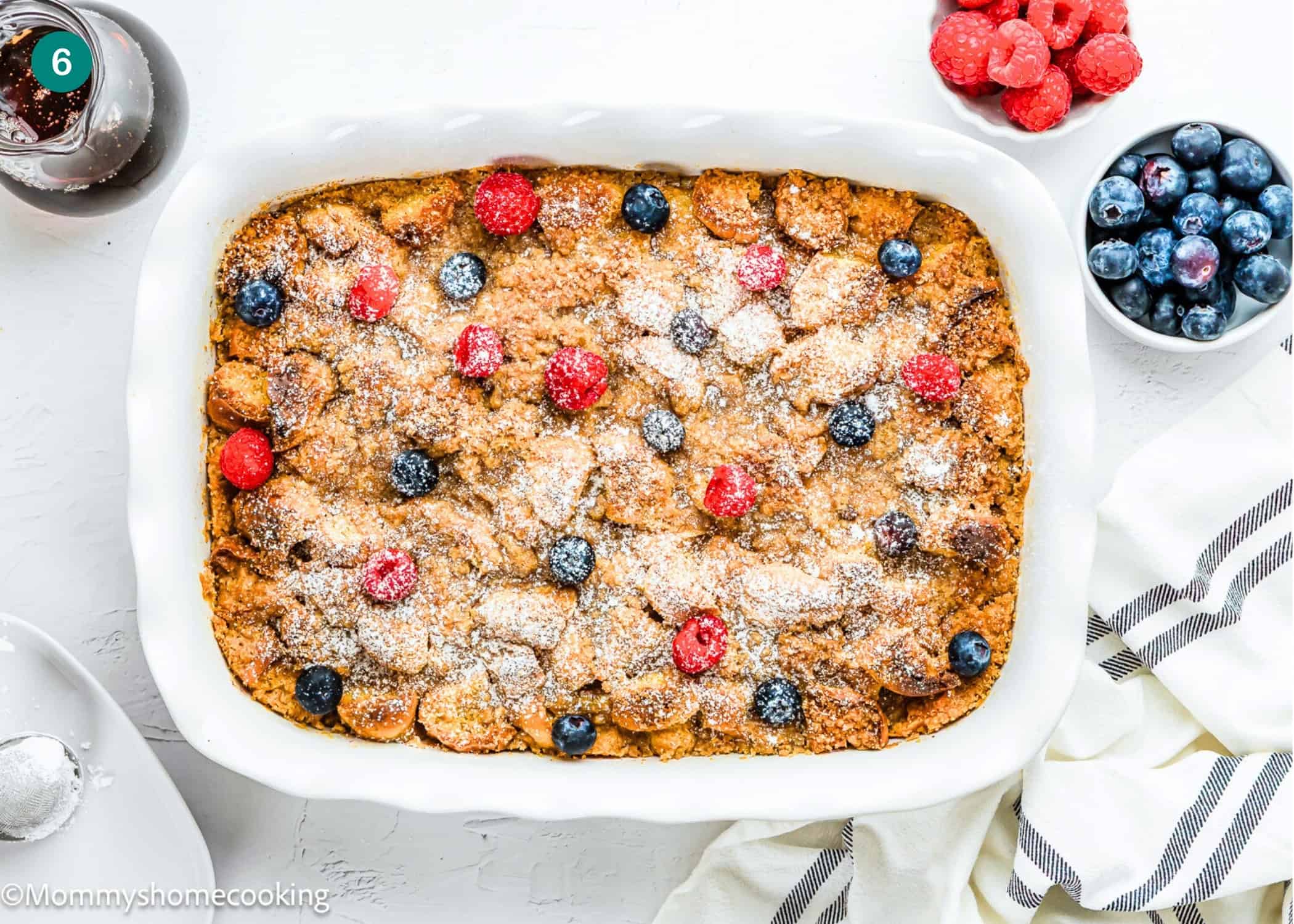 Overnight French Toast Casserole Without Eggs baked to perfection, topped with powdered sugar, raspberries, and blueberries in a white dish, surrounded by bowls of fresh berries and a striped towel.