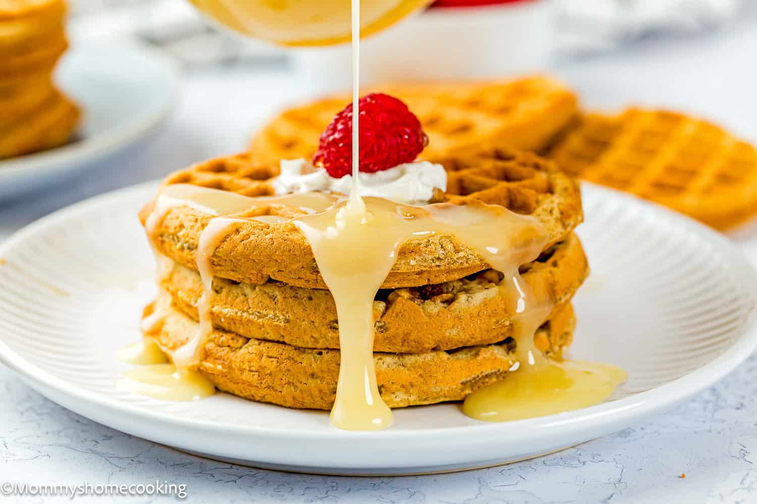 Three stacked waffles topped with whipped cream and a raspberry are being drizzled with an easy cream cheese glaze on a white plate.