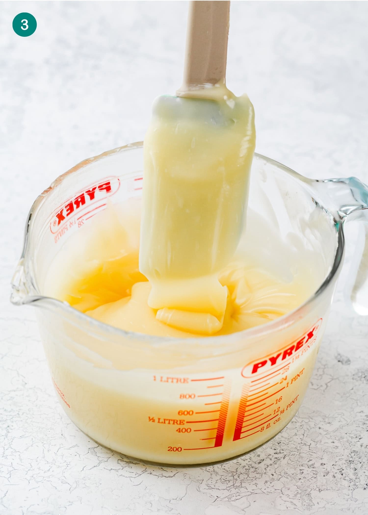 A spatula coated with thick yellow batter rests inside a Pyrex measuring cup filled with the same mixture, ready to be topped with Easy Cream Cheese Glaze, on a white countertop.