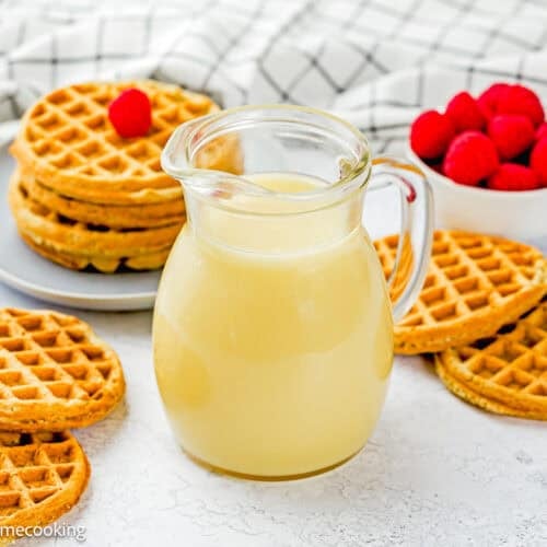 A glass pitcher of light syrup sits on a white surface, surrounded by stacks of waffles, a bowl of fresh raspberries, and an easy cream cheese glaze.