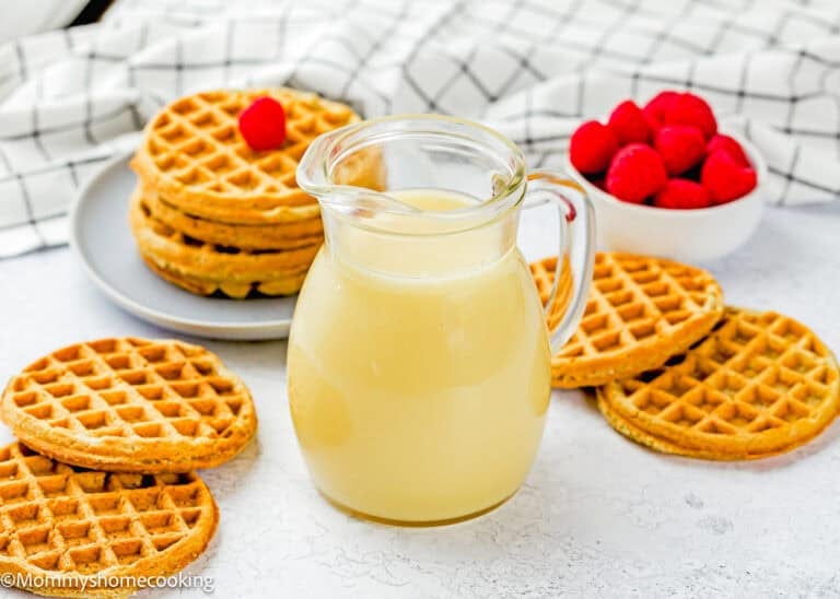A glass pitcher of light syrup sits on a white surface, surrounded by stacks of waffles, a bowl of fresh raspberries, and an easy cream cheese glaze.