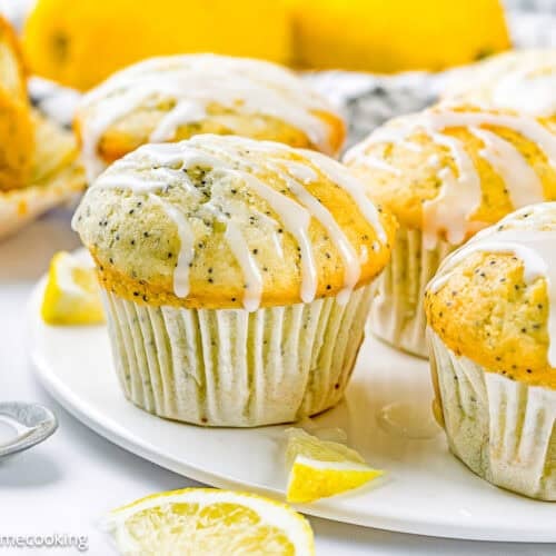 Egg-Free Lemon Poppy Seed Muffins with a white glaze drizzle are arranged on a white plate, surrounded by lemon slices and a bowl of glaze.