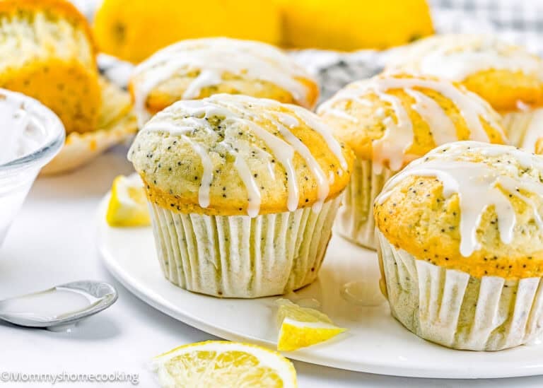 Egg-Free Lemon Poppy Seed Muffins with a white glaze drizzle are arranged on a white plate, surrounded by lemon slices and a bowl of glaze.