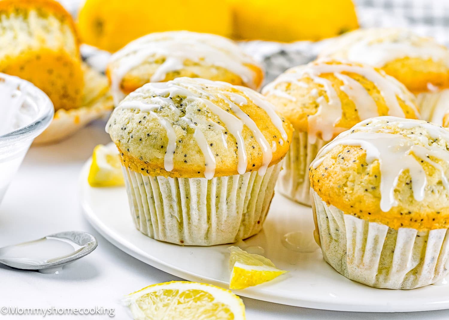 Egg-Free Lemon Poppy Seed Muffins with a white glaze drizzle are arranged on a white plate, surrounded by lemon slices and a bowl of glaze.