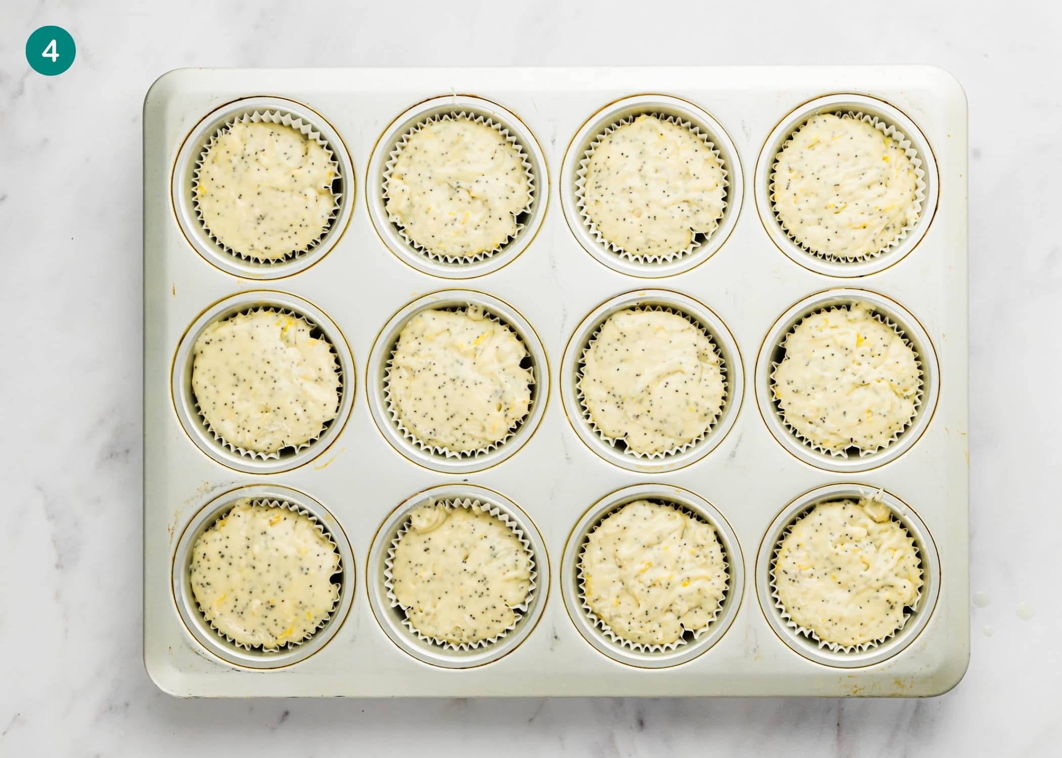 A muffin tin filled with 12 paper liners, each containing unbaked Egg-Free Lemon Poppy Seed Muffins batter with visible poppy seeds, sits on a white marble countertop.