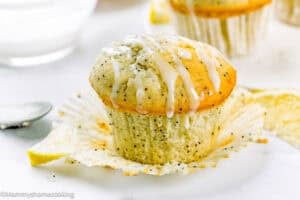 An Egg-Free Lemon Poppy Seed Muffin with white icing drizzle sits on a partially unwrapped paper liner. Lemon slices are visible in the background.