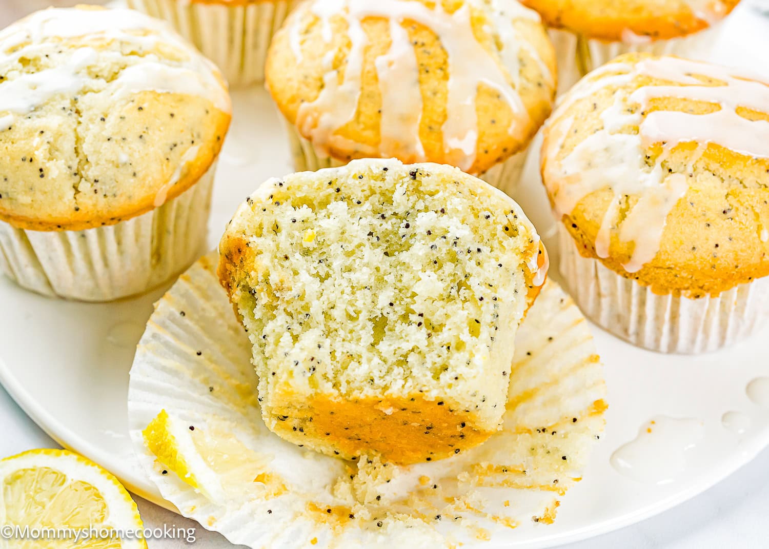A plate of Egg-Free Lemon Poppy Seed Muffins, one cut in half to show the moist interior, with icing drizzled on top.
