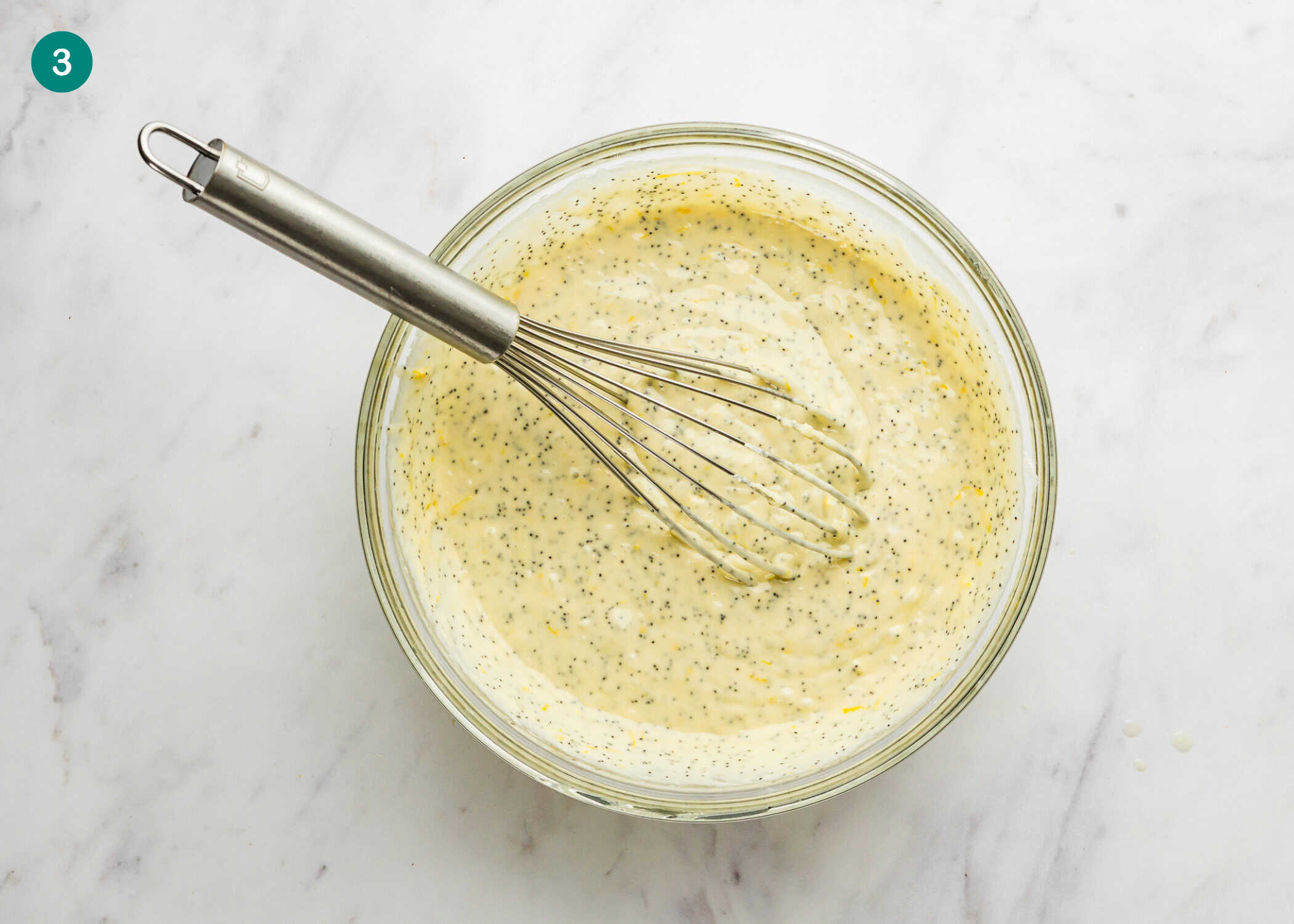 A glass bowl filled with batter for Egg-Free Lemon Poppy Seed Muffins, speckled with poppy seeds and mixed with a metal whisk, sits on a white marble surface.