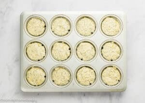 A 12-cup muffin pan filled with Egg-Free Lemon Poppy Seed Muffins batter in paper liners, viewed from above on a white marble surface.