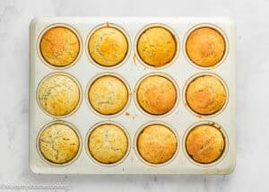 A muffin tray filled with twelve evenly baked Egg-Free Lemon Poppy Seed Muffins, some with visible poppy seeds, on a white surface.
