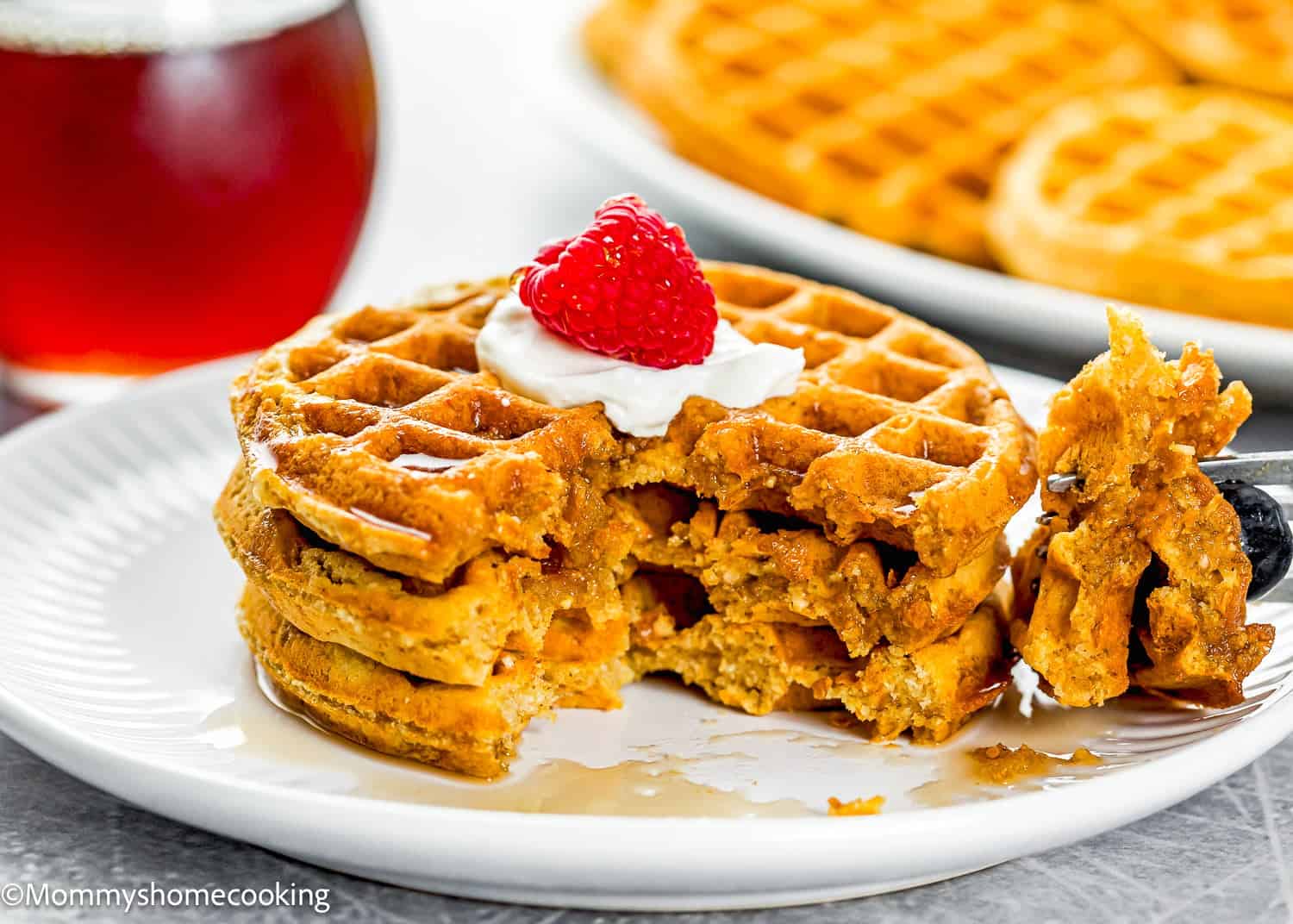 A stack of dairy-free and egg-free oat waffles with a bite taken out, topped with whipped cream and a raspberry, served on a white plate with a glass of syrup in the background.
