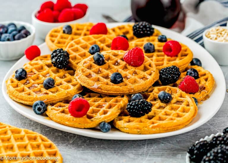 A plate of egg-free and refined sugar–free oat waffles topped with fresh raspberries, blueberries, and blackberries sits in front of bowls filled with berries.