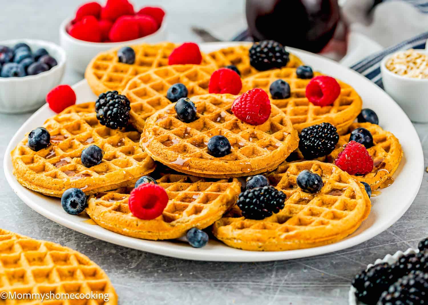 A plate of egg-free and refined sugar–free oat waffles topped with fresh raspberries, blueberries, and blackberries sits in front of bowls filled with berries.