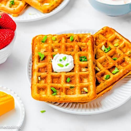 Two square Eggless Bacon Cheddar Savory Waffles topped with sour cream and chopped green onions on a white plate, with a bowl of strawberries and a block of cheddar cheese nearby.