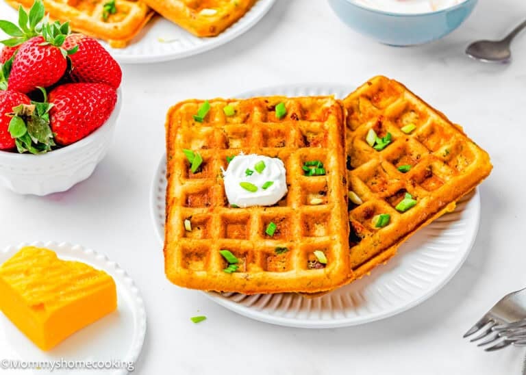 Two square Eggless Bacon Cheddar Savory Waffles topped with sour cream and chopped green onions on a white plate, with a bowl of strawberries and a block of cheddar cheese nearby.