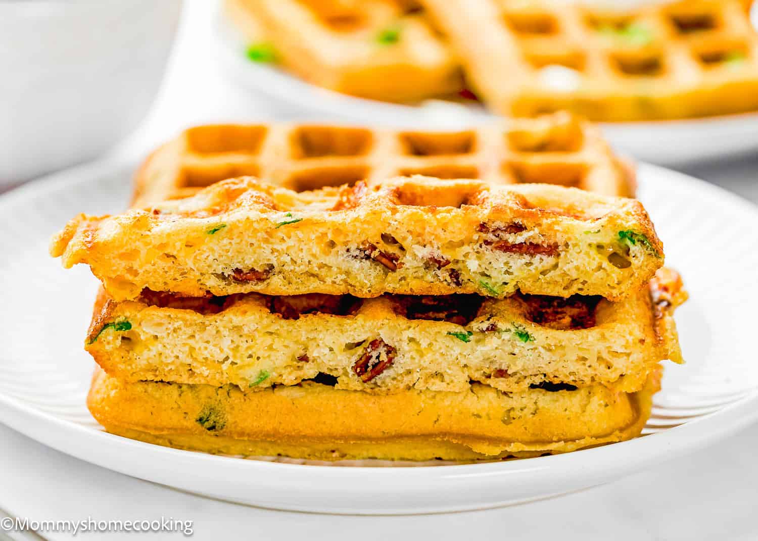 Two stacked Eggless Bacon Cheddar Savory Waffles with visible pieces of bacon and green herbs served on a white plate.