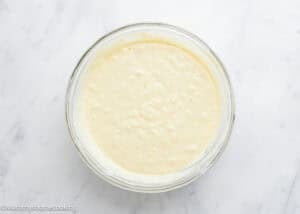 A glass bowl filled with batter for Eggless Bacon Cheddar Savory Waffles sits on a white marble surface.