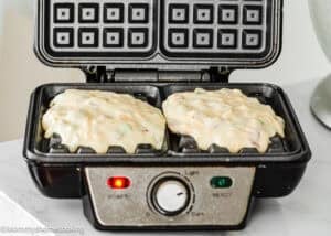 Batter for Eggless Bacon Cheddar Savory Waffles is poured into the compartments of an open waffle maker, which is turned on and ready to cook.