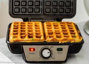 A waffle maker with two Eggless Bacon Cheddar Savory Waffles inside sits on a countertop, with power and ready indicator lights on the front panel.