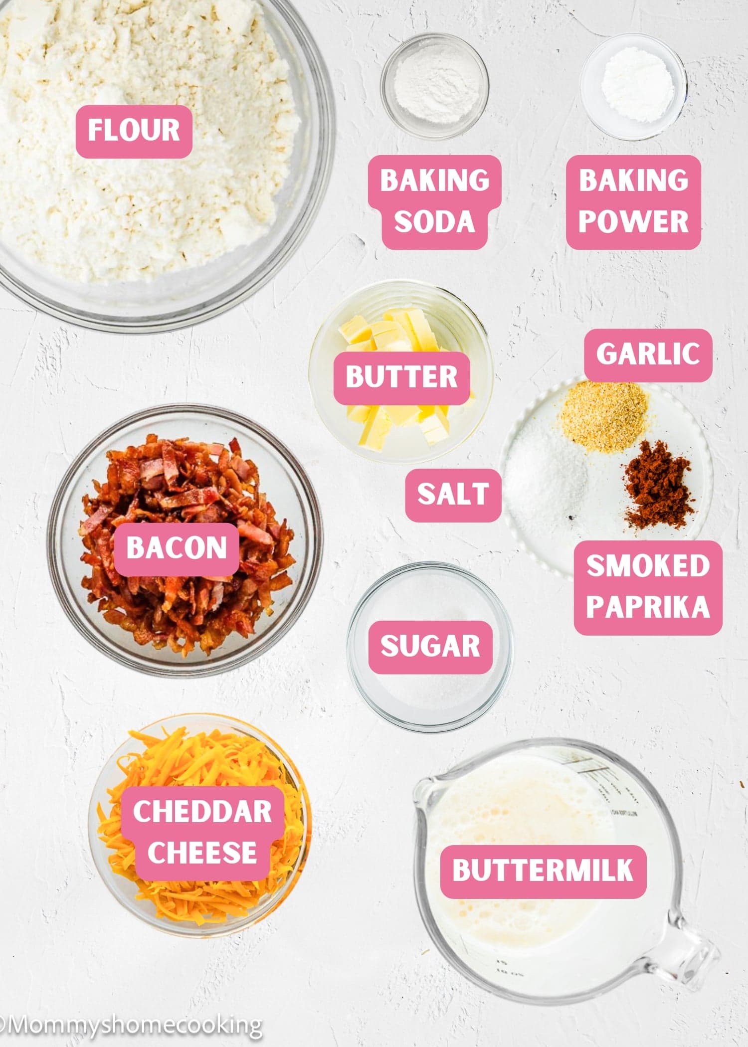 Top-down view of labeled baking ingredients for Eggless Bacon Cheddar Savory Waffles in bowls and containers, including flour, baking soda, baking powder, butter, salt, garlic, smoked paprika, bacon, sugar, cheddar cheese, and buttermilk.