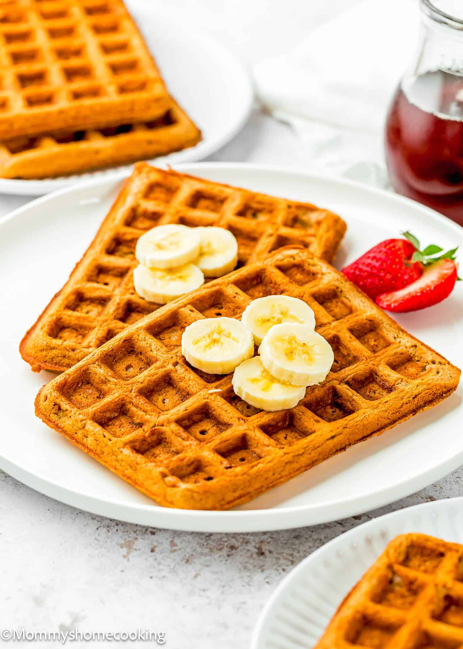 Two square Eggless Banana Waffles topped with banana slices are served on a white plate with a strawberry on the side; a small jar of syrup is in the background.