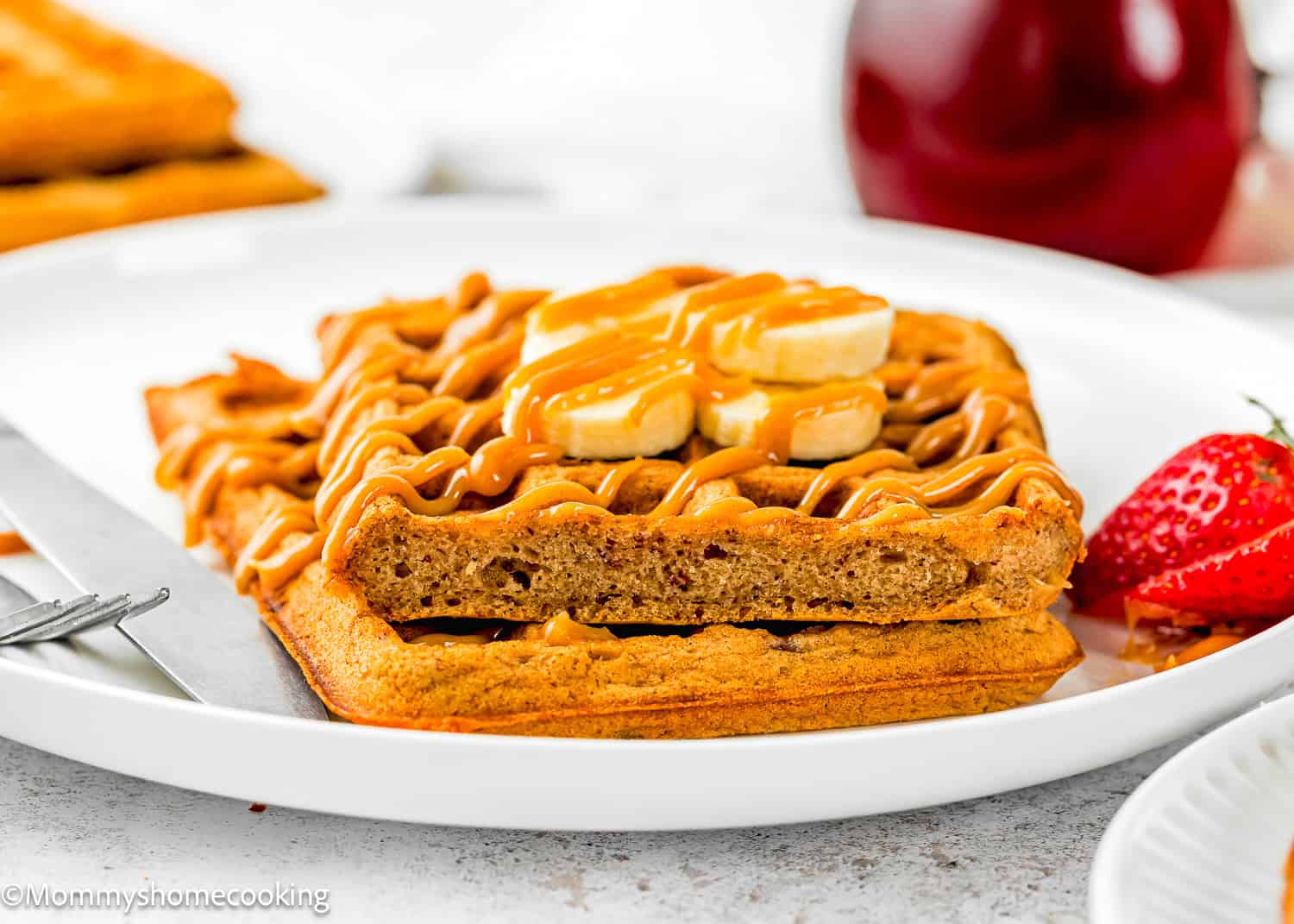 A plate with two stacked Eggless Banana Waffles topped with banana slices and drizzled sauce, served with a strawberry and a knife.