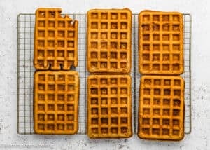 Six rectangular Eggless Banana Waffles cool on a wire rack; the top left waffle has two bites missing from one corner.