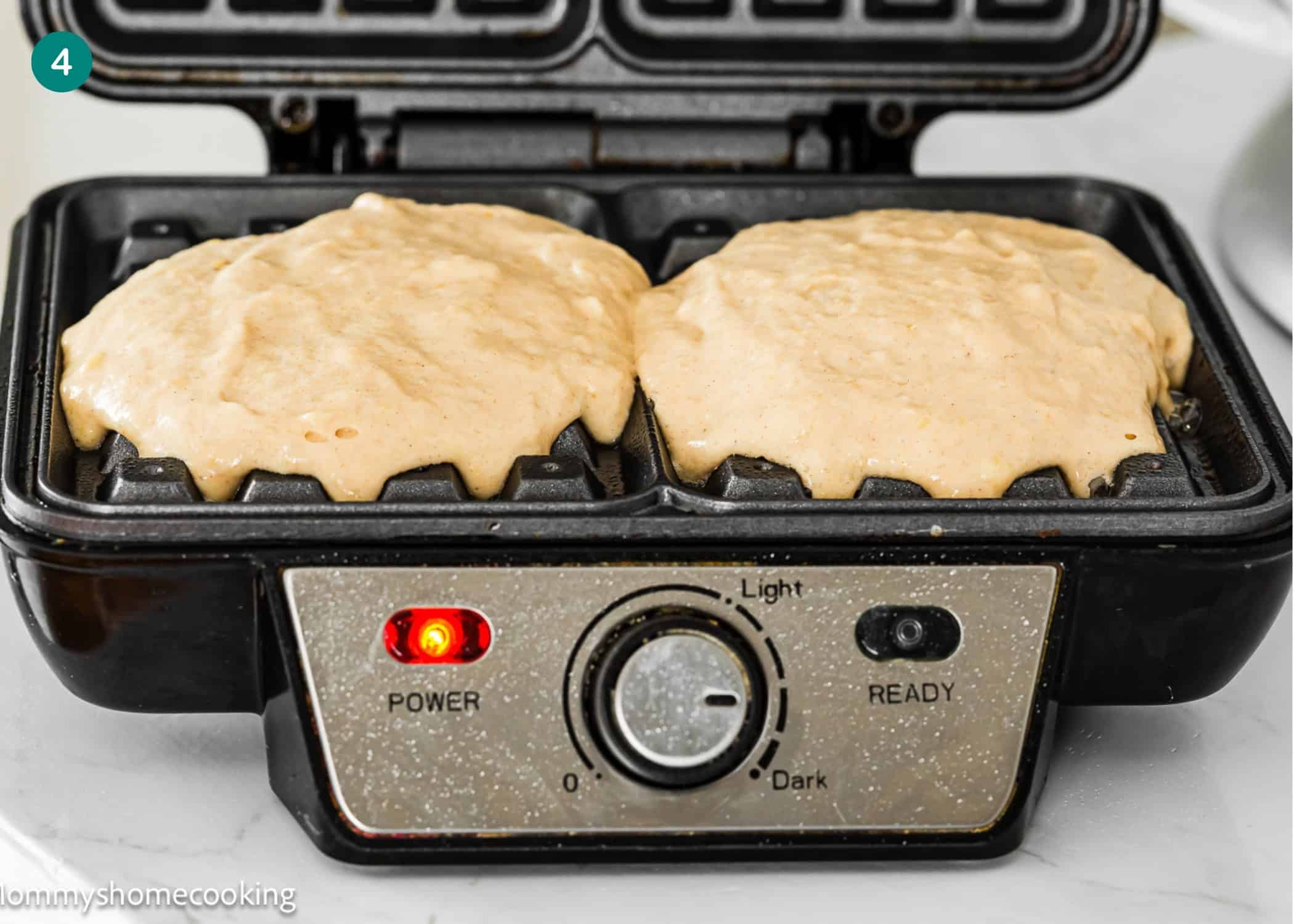 Close-up of a waffle maker with two portions of raw Eggless Banana Waffles batter inside, the power light on, and the control dial set between light and dark.