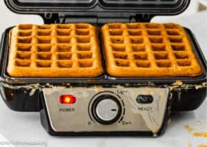 A waffle maker with two golden brown Eggless Banana Waffles inside, some batter overflow visible on the edges, and the power and light indicators turned on.