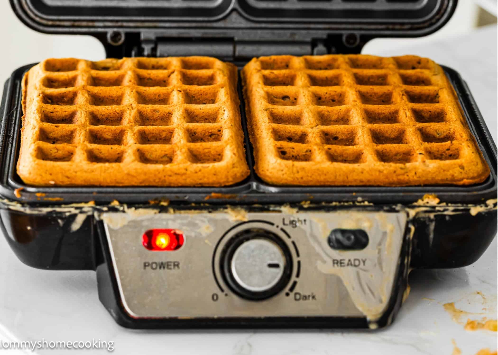 A waffle maker with two golden brown Eggless Banana Waffles inside, some batter overflow visible on the edges, and the power and light indicators turned on.