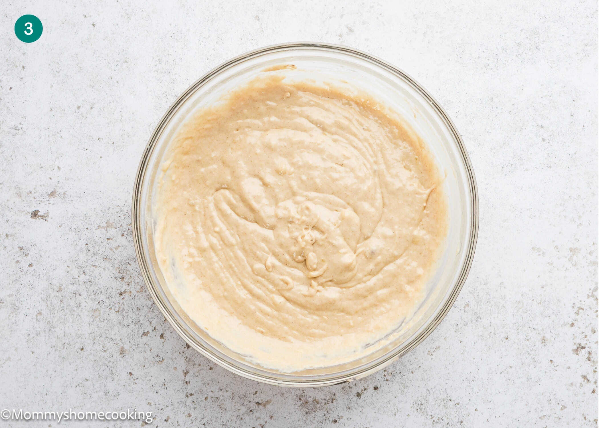 A clear glass bowl filled with smooth, light brown eggless banana waffles batter sits on a white textured surface.