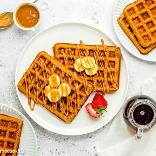 A plate of Eggless Banana Waffles topped with banana slices and caramel drizzle, served with a strawberry, syrup, and a side of caramel sauce. Extra waffles are on separate plates.