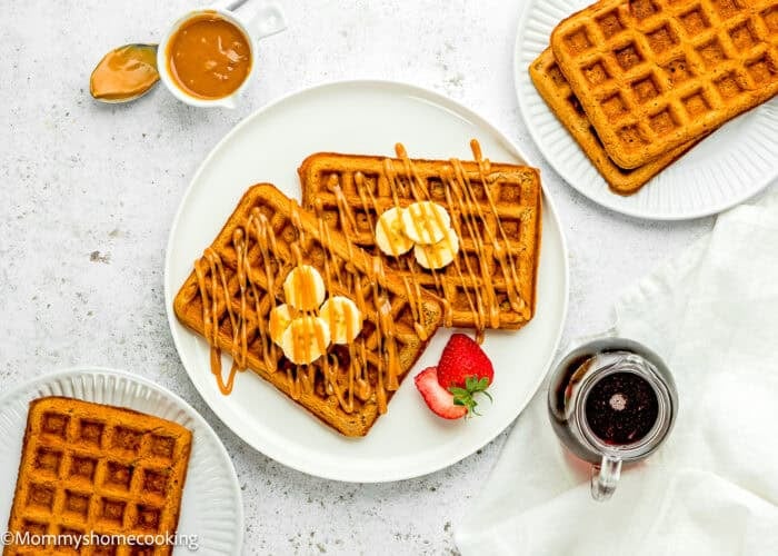 A plate of Eggless Banana Waffles topped with banana slices and caramel drizzle, served with a strawberry, syrup, and a side of caramel sauce. Extra waffles are on separate plates.