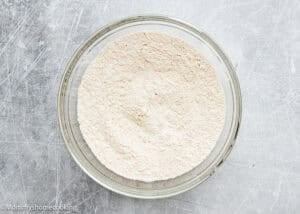 A glass bowl filled with a mound of dry flour mixture for Eggless Carrot Bundt Cake sits on a grey textured surface.